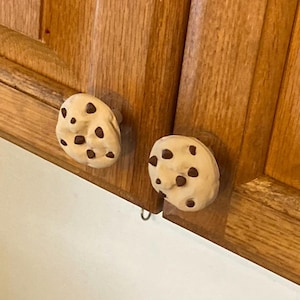 May include: Two round cabinet knobs shaped like beige chocolate chip cookies with dark brown chocolate chip details. The knobs are attached to wooden cabinet doors with a natural grain finish, providing a functional and textured appearance.