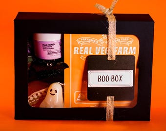 May include: A black gift box with a clear window, containing a jar of nighttime skin cream, a bat decoration, a ghost candle, and a package labeled "Real Veg Farm." A black tag with "Boo Box" is tied to the box.