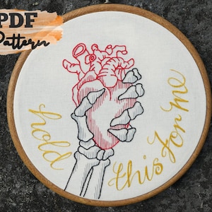 May include: Embroidered hoop art featuring a skeletal hand holding a red anatomical heart. The text "hold this for me" is embroidered in gold thread.