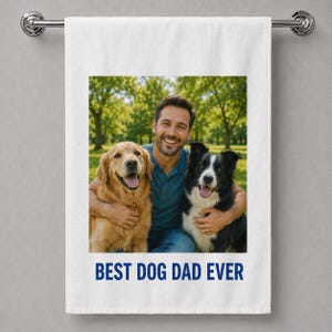 May include: A white rectangular towel featuring a printed photograph of a person with a golden retriever and a black and white border collie. Below the image, the text BEST DOG DAD EVER is printed in bold blue capital letters.