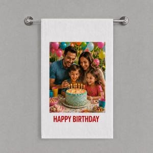May include: A white rectangular towel hanging on a silver metal rod, featuring a printed photograph of a family celebrating a birthday with a lit cake, colourful balloons, and the red text HAPPY BIRTHDAY printed at the bottom of the towel.