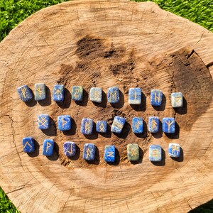 May include: A set of 25 blue lapis lazuli rune stones with gold lettering on a wooden surface. Each stone features a different rune symbol.