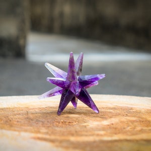 Könnte beinhalten: Ein violetter Amethyst-Kristallstern, auch bekannt als Merkaba, liegt auf einer Holzoberfläche. Der Kristall ist facettiert und hat eine spitze Form.