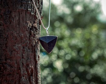 Peut inclure: Un collier composé d'un pendentif triangulaire sombre à la surface facettée, suspendu à une fine chaîne argentée. Le pendentif présente une texture mate et lisse, maintenu par une bélière métallique fixée à la chaîne.