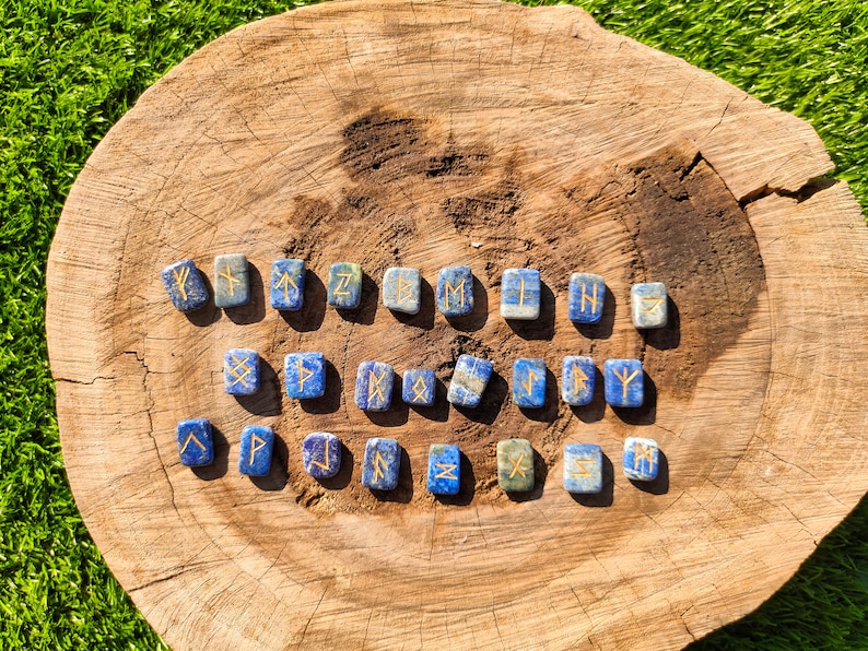 May include: A set of 24 lapis lazuli rune stones with gold lettering on a wooden surface. Each stone is a square shape and features a different rune symbol.