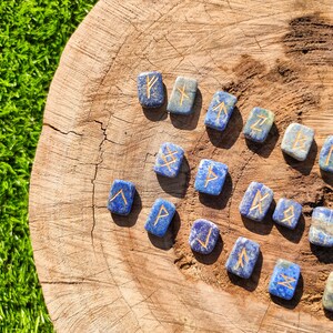 May include: A set of blue and gold rune stones on a wooden surface. The runes are carved into the stones and are surrounded by a light brown background.