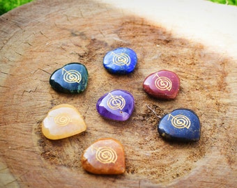 Puede incluir: Siete piedras en forma de corazón con símbolos dorados sobre una superficie de madera. Las piedras son de los colores de los chakras: rojo, naranja, amarillo, verde, azul, índigo y violeta.