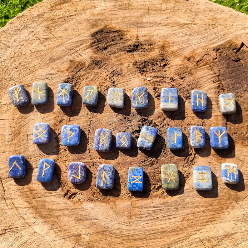 May include: A set of 24 blue lapis lazuli stones with gold runes engraved on them. The stones are arranged in rows on a wooden surface.