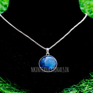 Puede incluir: Un collar de plata con un colgante redondo azul. El colgante tiene un diseño veteado azul oscuro y azul claro. El collar se muestra sobre un fondo negro. El texto "NKINTERNATIONALSIN" es visible.