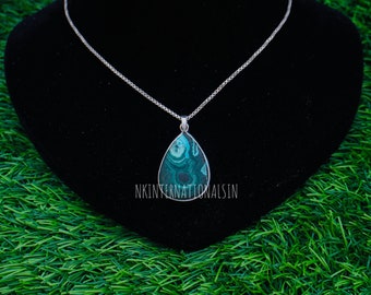 May include: A silver necklace with a teardrop-shaped pendant. The pendant features a teal and dark green stone with a unique pattern. The necklace is displayed on a black velvet surface against a green background. The text "NKINTERNATIONALSIN" is visible.