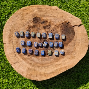 May include: A set of 20 blue lapis lazuli stone runes with gold lettering on a wooden surface. Each rune is a different symbol from the Elder Futhark.