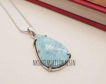 May include: A teardrop-shaped pendant with a light blue gemstone, set in a silver-colored metal frame, hangs from a silver chain. The text "NKINTERNATIONALSIN" is visible on the pendant. The background includes a red-covered book.