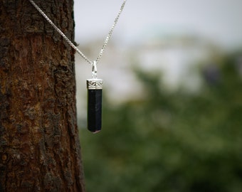 Puede incluir: Un collar de cadena de plata con un colgante de piedra negra. El colgante tiene una forma rectangular alargada con un borde plateado.