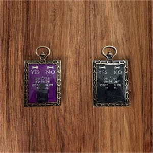 May include: Two rectangular pendants with the words "YES" and "NO" in white lettering. One pendant has a purple background, the other a black background. Both have a circuit board design and are framed in a bronze-colored metal with a loop at the top.