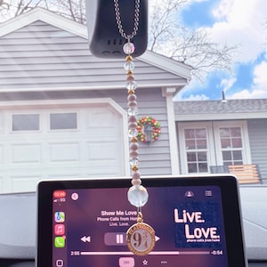 May include: A car charm featuring a gold-colored pendant with the number 9 3/4, surrounded by clear and pink beads. The charm hangs from a silver chain. The text "Live. Love." is visible on a blue background.