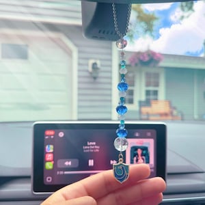 May include: A car charm featuring a beaded strand with blue, white, and clear beads. A small, blue and silver shield charm hangs at the bottom. The charm is suspended in front of a car's dashboard.