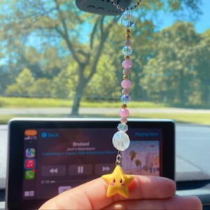 May include: A yellow star-shaped car charm with a smiling face hangs from a beaded chain with pink and clear beads. The charm is hanging from the rearview mirror of a car.