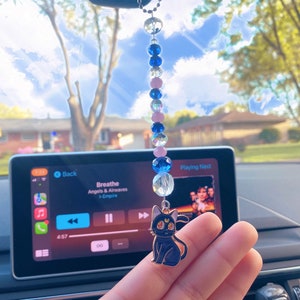 May include: A black cat charm with a crescent moon on its forehead hangs from a rearview mirror. The charm is attached to a beaded chain with pink, blue, and clear beads. The car's infotainment screen displays the song "Breathe" by Angels & Airwaves.