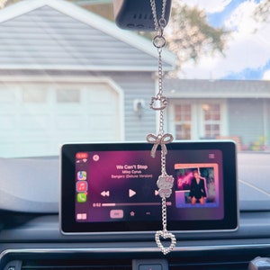 May include: A silver car charm with a Hello Kitty character, a bow, and a heart hanging from the rearview mirror of a car. The car's infotainment system is playing the song "We Can't Stop" by Miley Cyrus.