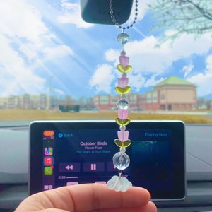 May include: A pink and yellow beaded car charm with a cloud-shaped charm hanging from it. The charm is hanging from the rearview mirror of a car. The car's infotainment system is visible in the background, playing music by October Birds.