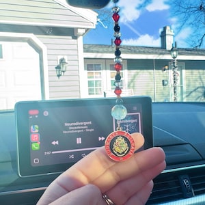 May include: A gold-colored car charm featuring the Hogwarts crest with red, yellow, and black accents. The charm is hanging from a rearview mirror in a car. The car's infotainment system is playing the song "Neurodivergent" by Stayfelmusic.