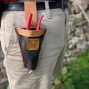 May include: A black leather tool holder with a brown leather flap and a metal buckle. The holder is attached to a brown belt and has two red pruning shears inside.