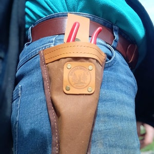 May include: A brown leather tool holder with a red and black pair of pruning shears. The holder is attached to a pair of blue jeans with a brown leather belt.