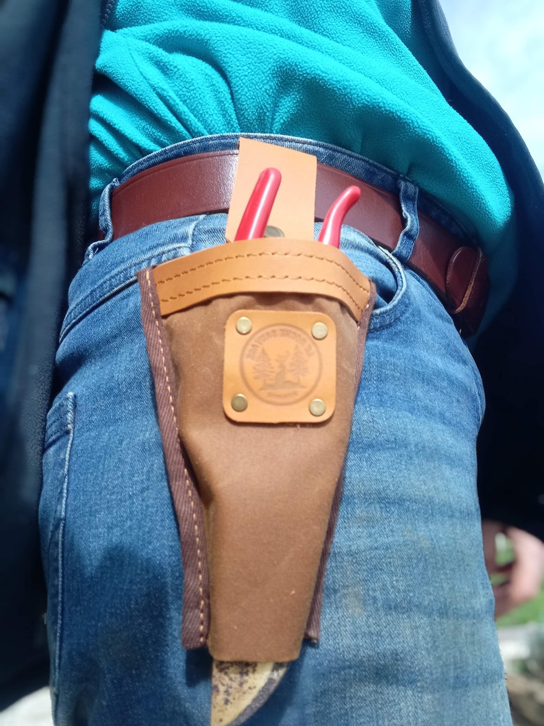 May include: A brown leather tool holder with a red and black pair of pruning shears. The holder is attached to a pair of blue jeans with a brown leather belt.