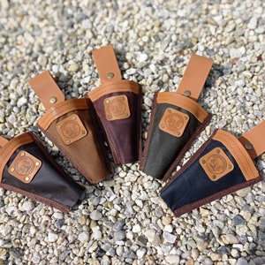 May include: Five different colored canvas tool holders with leather straps and a brown leather patch with a logo. The tool holders are arranged in a circle on a bed of small white rocks.
