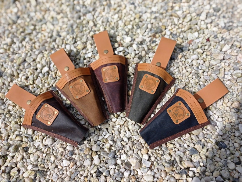 May include: Five different colored canvas tool holders with leather straps and a brown leather patch with a logo. The tool holders are arranged in a circle on a bed of small white rocks.