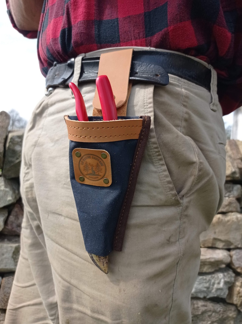 May include: A black canvas tool holder with a brown leather trim and a brown leather strap. The holder has a brass rivet and a logo that says "The Tree Trimmer". The holder is attached to a brown leather belt and is holding a pair of red pruning shears.