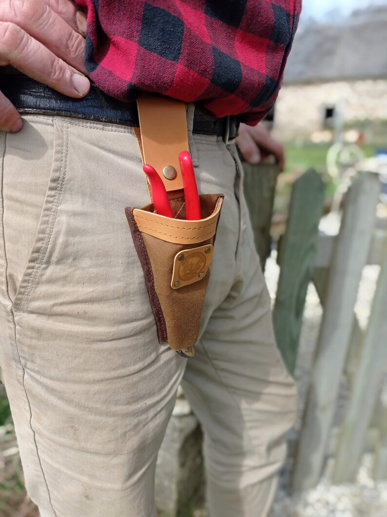 May include: A brown leather tool belt with a brown leather sheath for holding pruning shears. The shears are red and have a metal clasp. The belt is worn by a person wearing a red and black plaid shirt and beige pants.