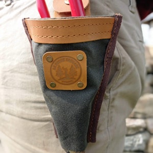 May include: A dark green canvas tool pouch with a brown leather flap and a brown leather strap. The pouch has a brown leather patch with the text "Le Rosier Sauvage" and a tree design. The pouch is attached to a belt.