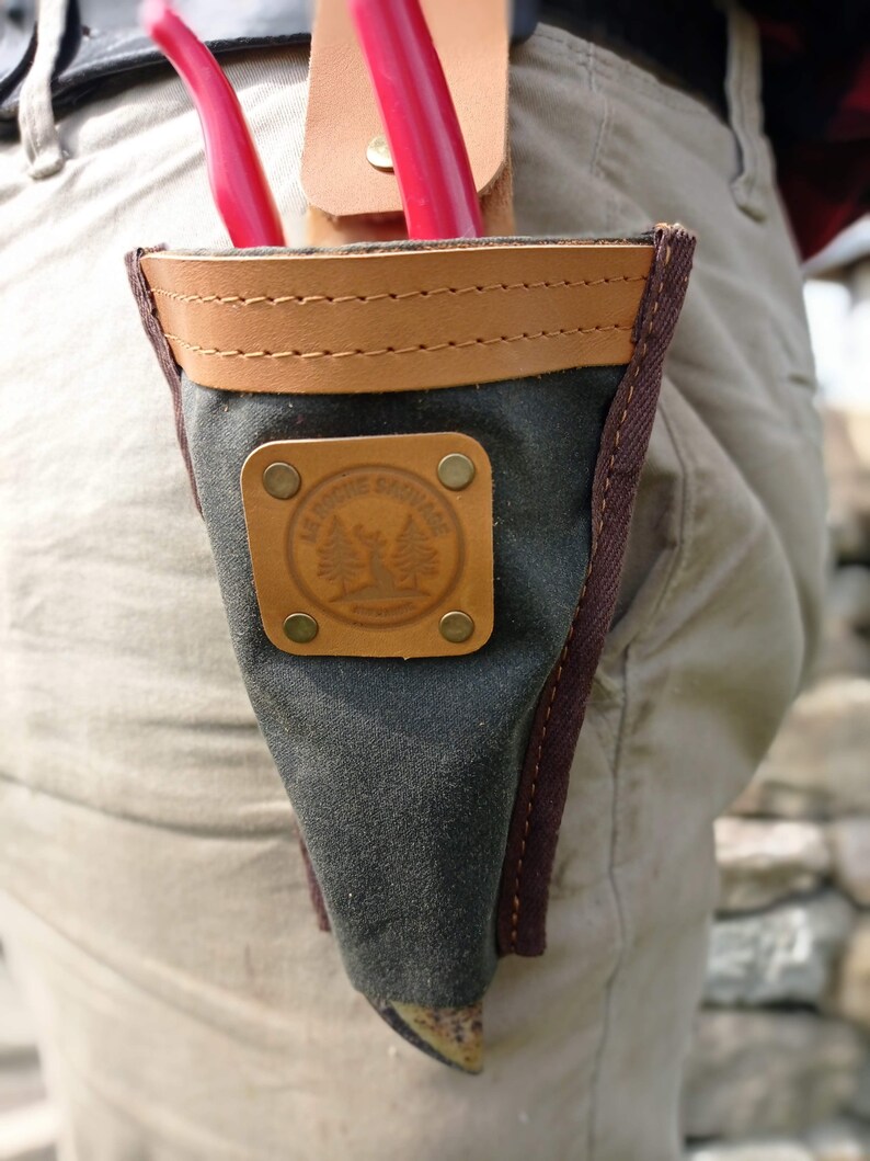 May include: A dark green canvas tool pouch with a brown leather flap and a brown leather strap. The pouch has a brown leather patch with the text "Le Rosier Sauvage" and a tree design. The pouch is attached to a belt.