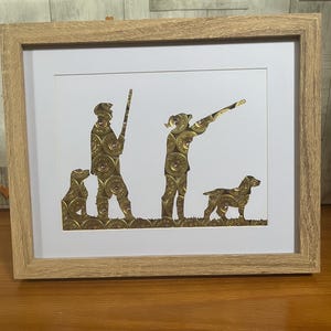 May include: A framed wall art piece featuring a silhouette of two figures and two dogs created from gold-coloured shotgun shell casings. The figures are positioned in a field setting, with one holding a rifle and the other aiming a shotgun.
