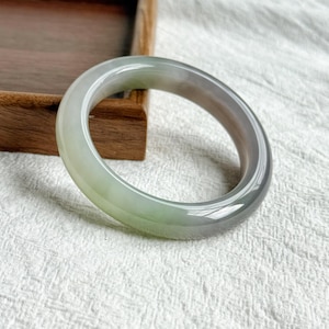 May include: A smooth, polished bangle bracelet crafted from translucent stone, featuring a gradient of soft pale green and muted grey tones. The circular jewellery piece has a rounded profile and a glossy finish that reflects light across its surface.