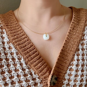 May include: A delicate gold necklace featuring a square, white pendant with a small green accent. The pendant hangs from a thin, gold chain. The necklace is displayed against a brown and white patterned sweater.