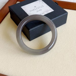 May include: A gray, translucent bangle bracelet. The smooth, circular bracelet is made of a polished stone. The bracelet is resting on a cream-colored surface, with a black box in the background that has the text "INKLOTUS Jewelry EST. 2022".