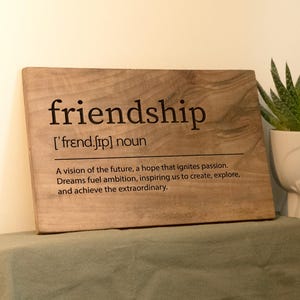 Könnte beinhalten: Ein Holzschild mit dem Wort "Freundschaft" definiert als "eine Vision der Zukunft, eine Hoffnung, die Leidenschaft entfacht. Träume befeuern den Ehrgeiz und inspirieren uns, zu erschaffen, zu erforschen und das Außergewöhnliche zu erreichen."