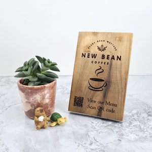Op de afbeelding: Houten bord met de tekst "Every Bean Matters" en "New Bean Coffee" met een gestileerde koffiekop en stoom. Het bord heeft ook een QR-code met de tekst "View our Menu Scan QR code".