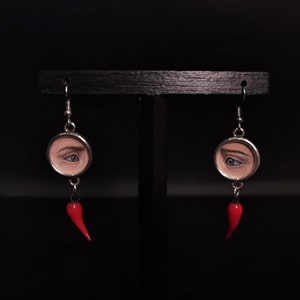 Puede incluir: Un par de pendientes de plata con un dije de chile rojo. Cada pendiente tiene un colgante redondo con un ojo pintado.