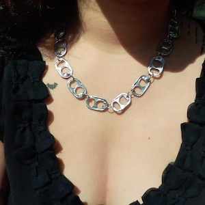 Może przedstawiać: Srebrny naszyjnik wykonany z przetworzonych zawleczek od puszek. Naszyjnik jest noszony na czarnym topie z falbankami. Wyjątkowy i przyciągający wzrok dodatek.