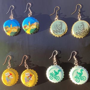 Könnte beinhalten: Handgefertigte Ohrringe aus recycelten Kronkorken. Die Ohrringe zeigen verschiedene Designs, darunter Landschaften, Bierlogos und ein Eichhörnchen auf einem Fahrrad. Die Kappen sind in Blau-, Gelb- und Grüntönen gehalten.