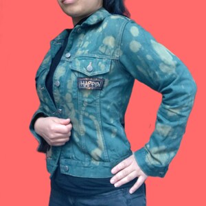 May include: Teal denim jacket with a bleached pattern and a small patch that reads "HAPPEN". The jacket has a classic collar, button closures, and two chest pockets. The jacket is worn over a black top and dark jeans.