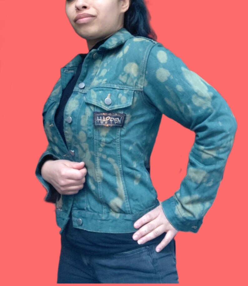 May include: Teal denim jacket with a bleached pattern and a small patch that reads "HAPPEN". The jacket has a classic collar, button closures, and two chest pockets. The jacket is worn over a black top and dark jeans.