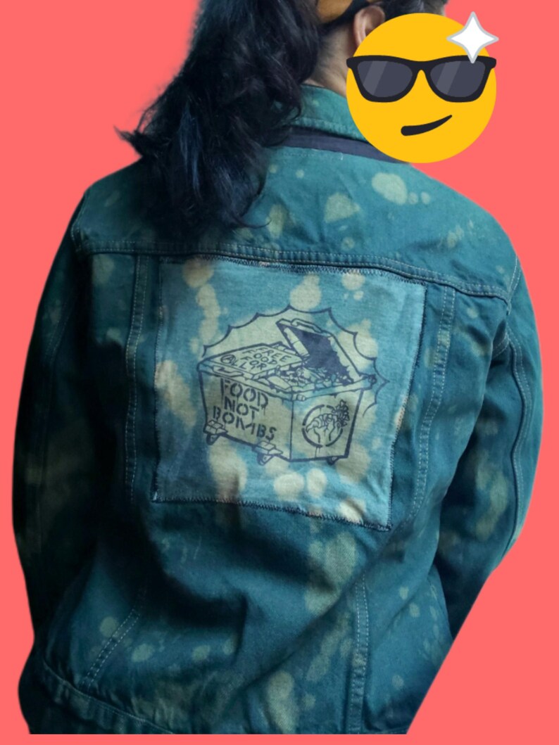 May include: Dark teal denim jacket with a bleached pattern. A patch on the back features a drawing of a dumpster overflowing with food, with the words "FOOD NOT BOMBS" and a fist symbol.