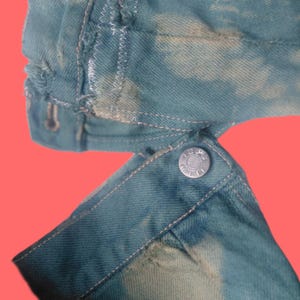 May include: Close-up of a teal denim jacket sleeve with a buttoned cuff. The denim has a bleached, cloud-like pattern. White stitching accents the seams and cuff. The jacket appears to be distressed.