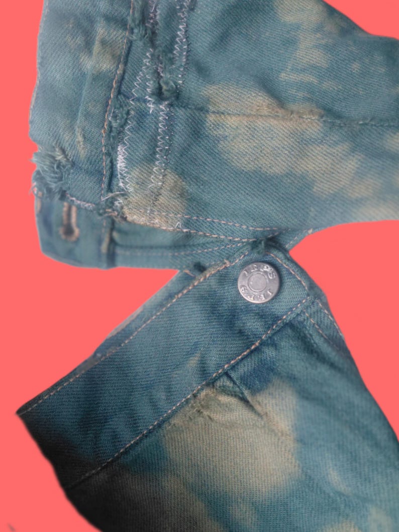 May include: Close-up of a teal denim jacket sleeve with a buttoned cuff. The denim has a bleached, cloud-like pattern. White stitching accents the seams and cuff. The jacket appears to be distressed.