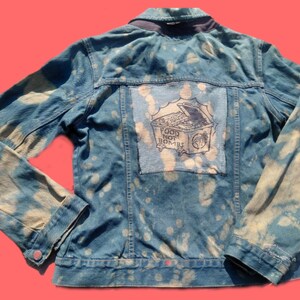 May include: Blue denim jacket with a bleached pattern. The back features a patch with the words "FOOD NOT BOMBS" and an illustration of a treasure chest. The jacket has long sleeves and a button closure.
