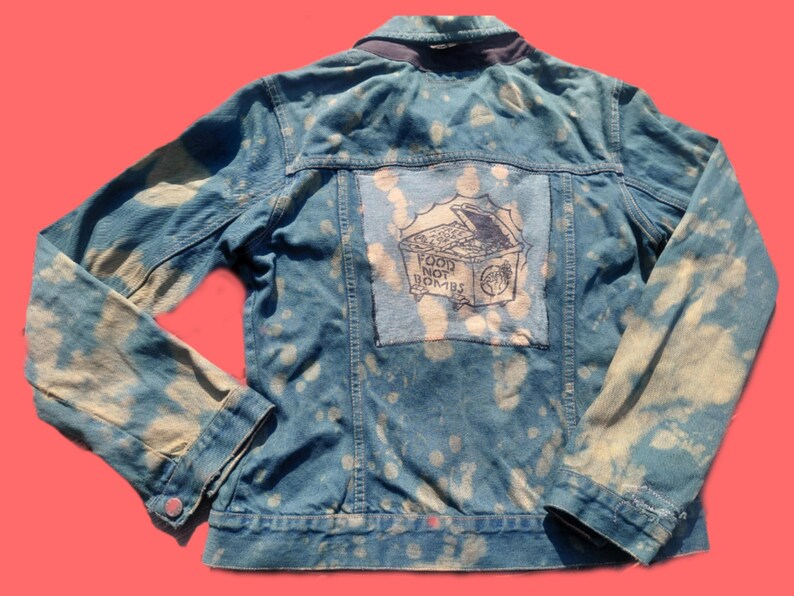 May include: Blue denim jacket with a bleached pattern. The back features a patch with the words "FOOD NOT BOMBS" and an illustration of a treasure chest. The jacket has long sleeves and a button closure.